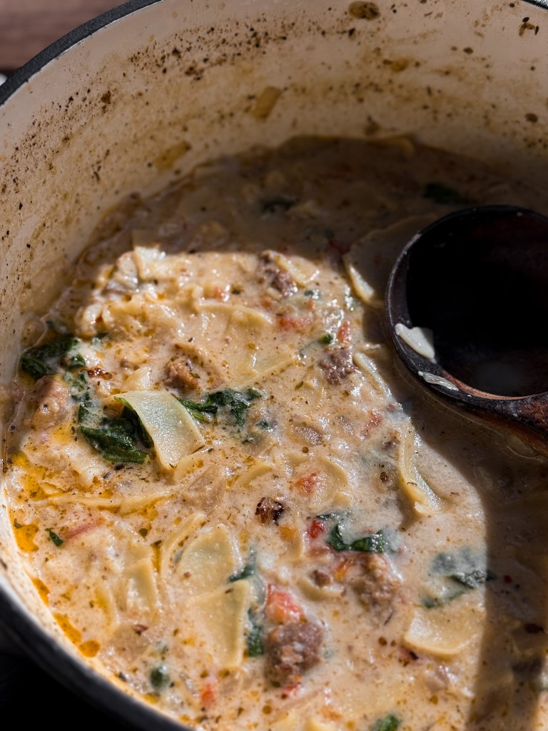 White Lasagna Soup in a Dutch Oven. Spinach, sun-dried tomatoes, tender noodles, and flavorful italian sausage and ground beef are peaking their way through the creamy and cheesy white broth.