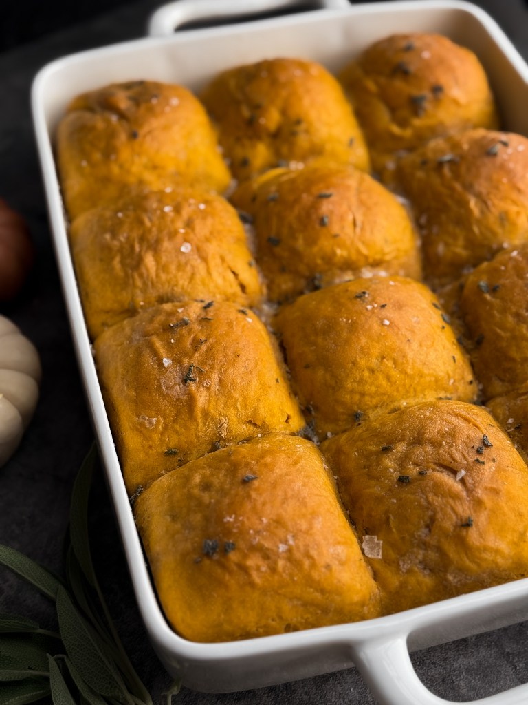 Pumpkin sage dinner rolls all lined up in a white 9x13 baking dish - all golden brown on top, smeared with fresh butter and sprinkled with chopped sage leaves and flaky sea salt. These Pumpkin Sage Dinner Rolls are a recipe by Christine Manes from Home-Cooked Living, a healthy recipe website.
