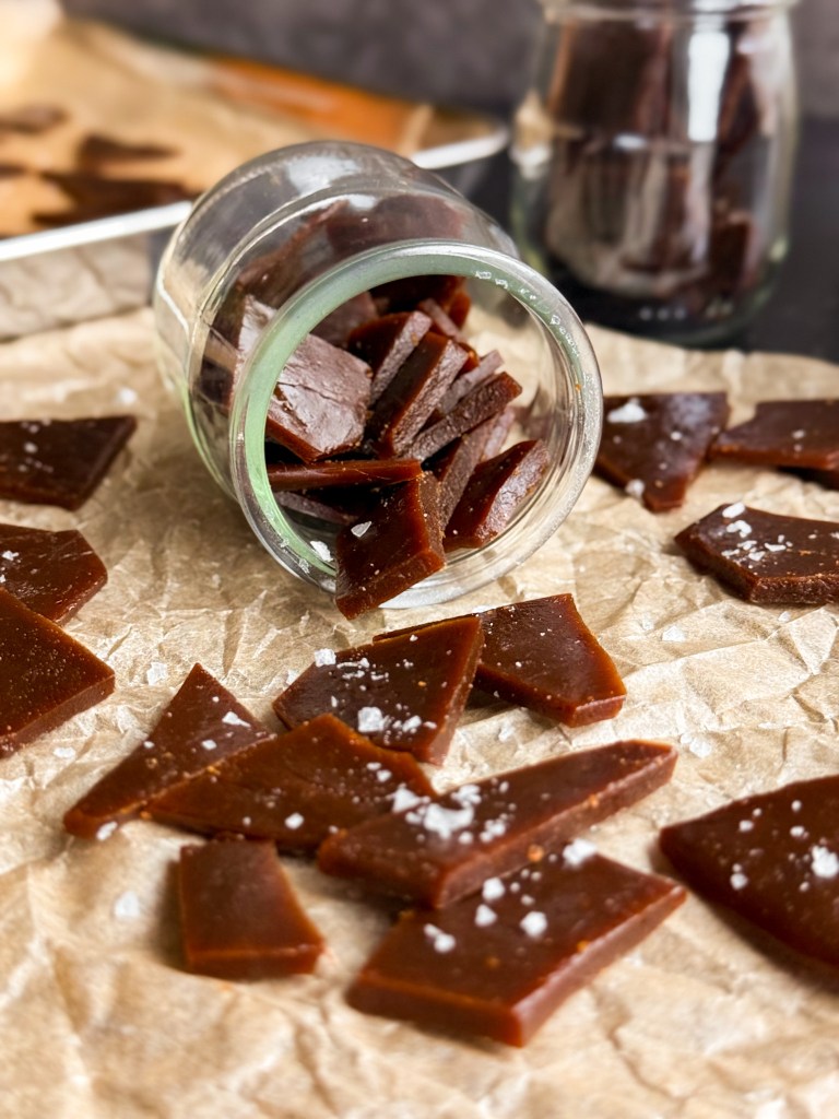 Toffee pieces falling out of a small jug onto parchment paper. This recipe is homemade toffee using coconut sugar as the sweetener - aka Coconut Sugar Toffee