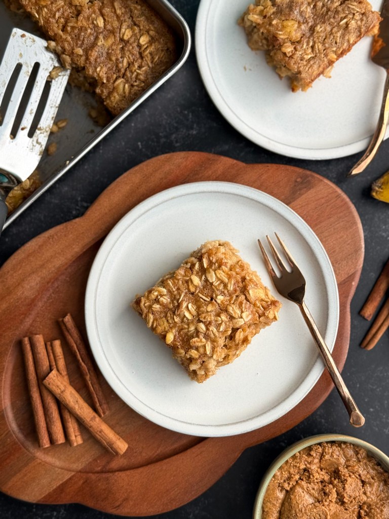 A hot slice of fresh baked Almond Butter Banana Baked Oatmeal on a white plate with a small metallic fork, all surrounded by cinnamon sticks for aesthetic