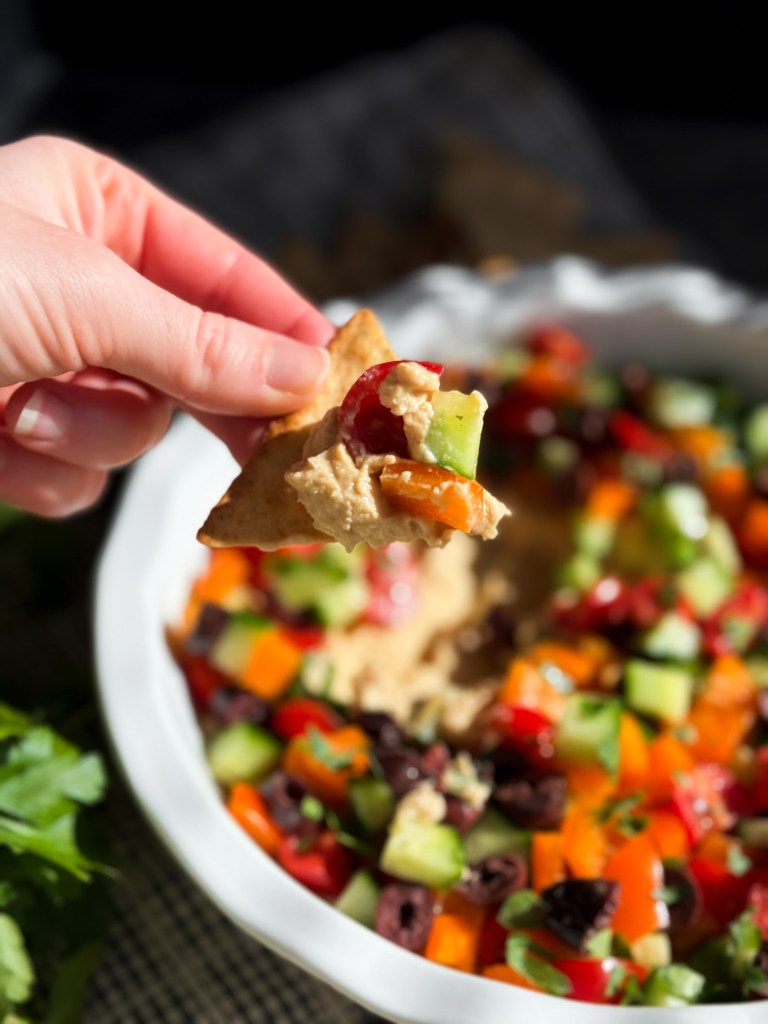 Pita cracker scooping Loaded Hummus with cucumber, cherry tomatoes, bell pepper, Kalamata olives, and fresh parsley.